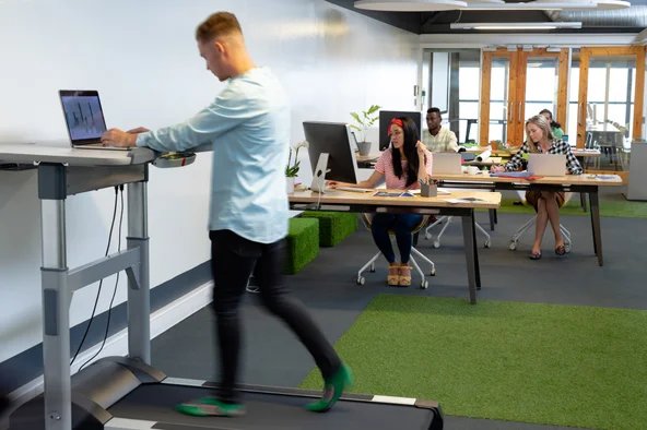 Ein Mitarbeiter arbeitet im Stehen an einem höhenverstellbaren Schreibtisch mit integriertem Laufband – modernes Büro als Beispiel für ergonomische Arbeitsplatzgestaltung und gelebten Arbeitsschutz.