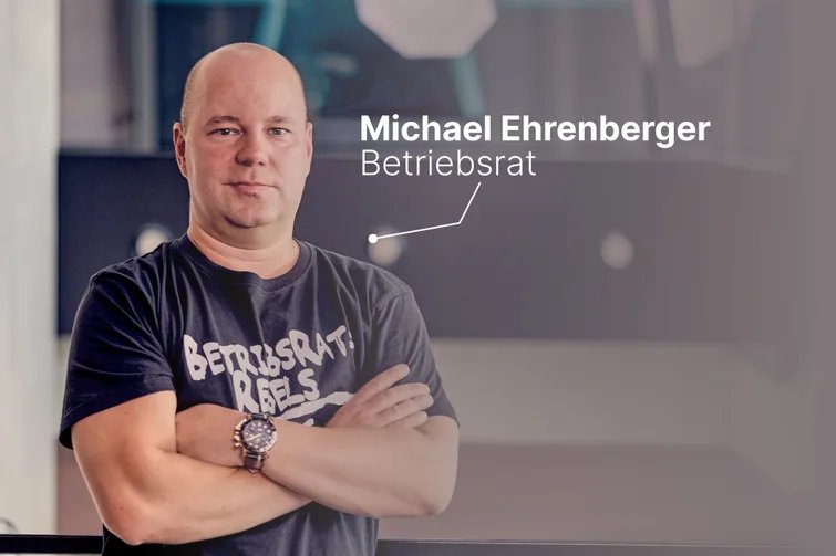 Ein Betriebsratsmitglied im T-Shirt mit der Aufschrift „Betriebsrat Rebels“ steht mit verschränkten Armen vor einem modernen Hintergrund – das Bild zeigt Michael Ehrenberger als engagierten und modernen Vertreter der Arbeitnehmerinteressen.